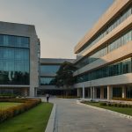 A modern exterior view of the Intel Technology India Pvt Ltd SRR1 facility in Bengaluru, featuring sleek glass buildings with the Intel logo, surrounded by lush greenery under a clear sky.