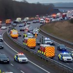 Police vehicles, ambulances, and fire trucks at the scene of a multi-vehicle accident on the closed M62 motorway near Huddersfield, with traffic backed up and emergency workers attending to the incident.