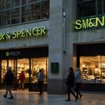 A brightly lit Marks and Spencer store with a prominent green sign, showcasing a busy shopping scene with customers entering and exiting, reflecting the retailer's recovery from a recent cyber attack.