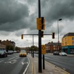 A busy urban intersection in Newham, East London, with a prominent yellow box junction marked on the road, monitored by an ANPR camera mounted on a pole under a cloudy sky.
