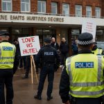 A group of protesters gathers outside a hotel used for housing asylum seekers in the UK, holding signs and banners, with police officers monitoring the scene under a cloudy sky.