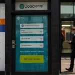 A person checks their bank account on a smartphone outside a Jobcentre Plus office, with a signboard in the background, symbolizing anticipation for rumored DWP £750 payments in June 2025.