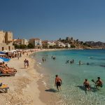 A vibrant beach scene on Rhodes, Greece, with British tourists enjoying the sunny shoreline, clear blue waters, and historic architecture in the background under a bright sky.