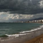 A view of La Carihuela beach in Benalmadena with murky brown water lapping at the shore, caused by a burst freshwater pipe, as concerned beachgoers stand at a distance under a cloudy sky.