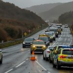 Emergency vehicles, including an ambulance and police cars, stationed along the A38 in Cornwall near Glynn Valley, with traffic cones and road closure signs visible under a cloudy sky.